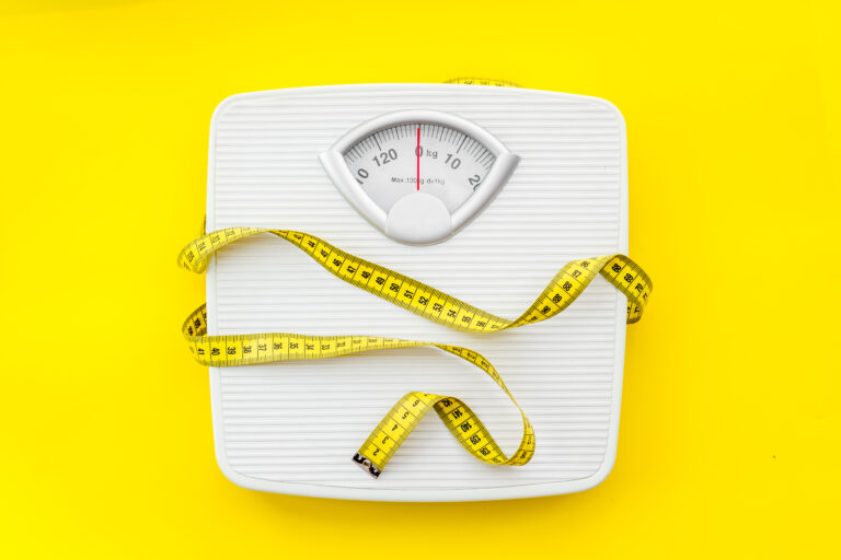 White bathroom scale with measuring tape wrapped around it on yellow background