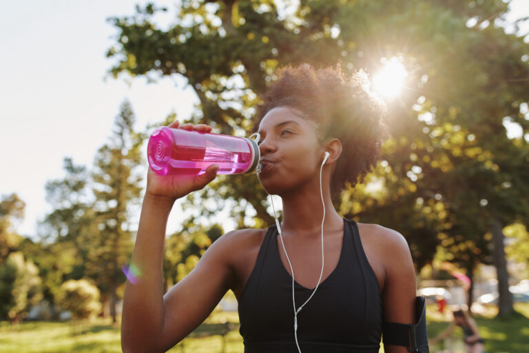 Woman drinking from pink water bottle outdoors during exercise with earbuds in