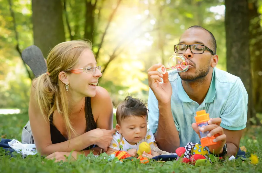 A mother, father and toddler lying on the grass in a sunny park, blowing bubbles