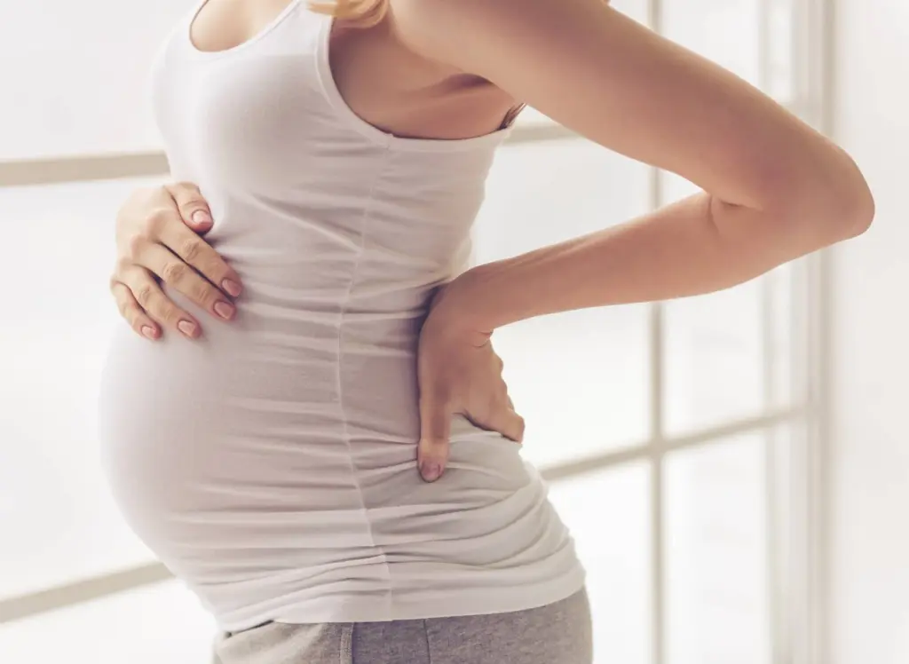 A pregnant woman standing by a bright window with one hand on her lower back