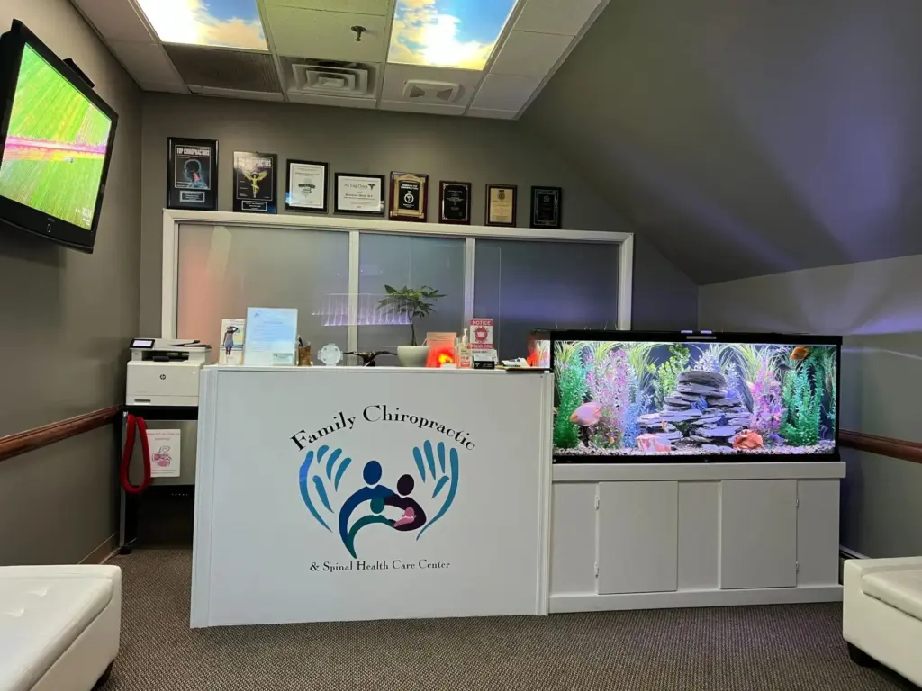 The front desk at Family Chiropractic in Ramsey: the practice's family-in-hands logo on the counter, a big planted aquarium beside it, award plaques on the wall and sky panels in the ceiling